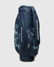 The OUTLINE BUNNY GOLF TOUR BAG - PBMG4FC1 by Psycho Bunny is a dark blue polyurethane golf cart bag with white branding. It features light blue embroidered bunny skull designs, multiple zippered compartments, and a detachable strap for convenience.