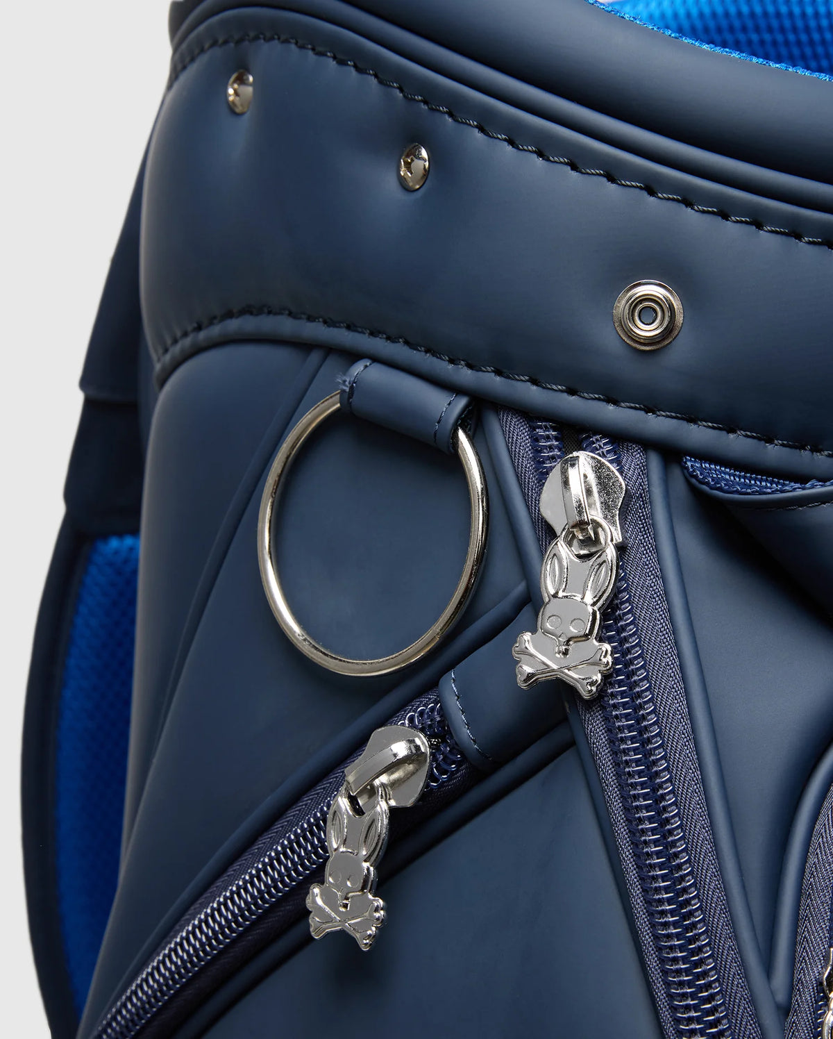 Close-up of Psycho Bunny's OUTLINE BUNNY GOLF TOUR BAG - PBMG4FC1, a blue polyurethane bag with silver skull and bunny zipper pulls. It resembles an elegant golf cart bag with a circular metal ring, visible stitching, detachable strap, and matching blue zippers and trim.