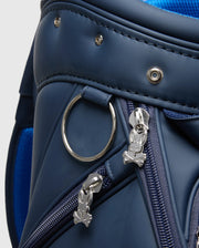 Close-up of Psycho Bunny's OUTLINE BUNNY GOLF TOUR BAG - PBMG4FC1, a blue polyurethane bag with silver skull and bunny zipper pulls. It resembles an elegant golf cart bag with a circular metal ring, visible stitching, detachable strap, and matching blue zippers and trim.