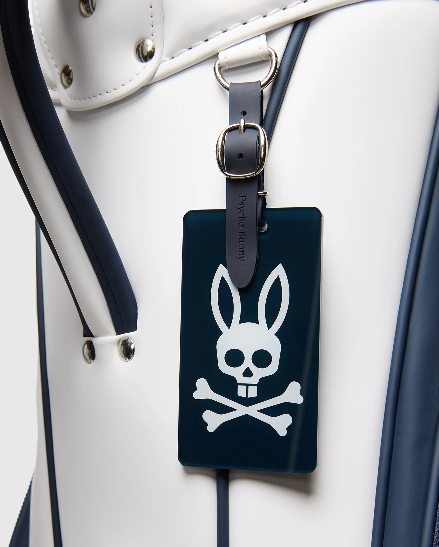 A close-up of the Psycho Bunny OUTLINE BUNNY GOLF TOUR BAG - PBMG4FC1 in white and dark blue, featuring a leather strap and a dark tag with a rabbit-eared skull and crossbones symbol.