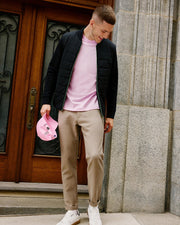 A person stands in front of a wooden door, wearing the Psycho Bunny MENS MYKEL MIXED MEDIA JACKET in black over a pink shirt, complemented by beige pants and white sneakers. They hold a pink cap in one hand and gaze downwards amidst a building with stone walls and decorative glass accents.