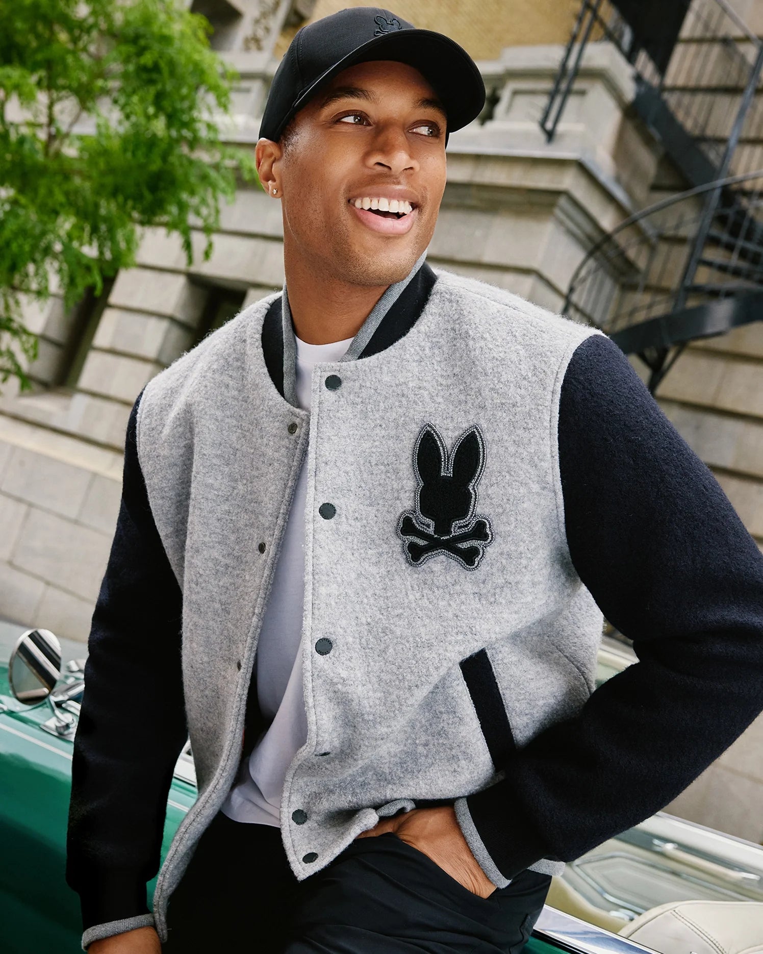 A person wearing a black cap and the stylish MENS TOM WOOL BOMBER JACKET - B6J744D200 from Psycho Bunny, adorned with a bunny patch, smiles while sitting on a car. In the background, there is a building with a fire escape surrounded by greenery.