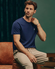 A man with a beard sits on a brown chair, dressed in the Psycho Bunny MENS CLASSIC CREW NECK TEE - B6U014CRPC and beige pants. The blue T-shirt, made of soft pima cotton, fits him perfectly in a regular fit. He gazes to the side with one hand near his face, set against a dark green curtain and wooden floor backdrop.