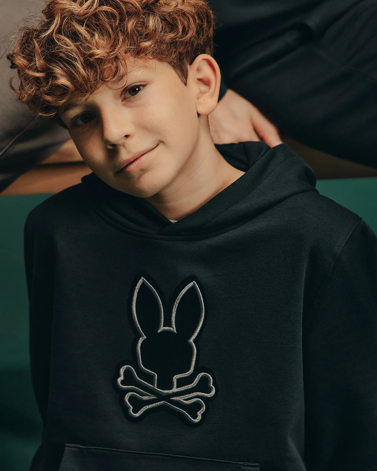 A boy with curly hair is wearing the Psycho Bunny Kids Alexander Hoodie (B0H154E200), which features a stylish bunny skull design. This versatile black hoodie perfectly complements his relaxed expression. The blurred background highlights his comfortable, laid-back fit and cool style.