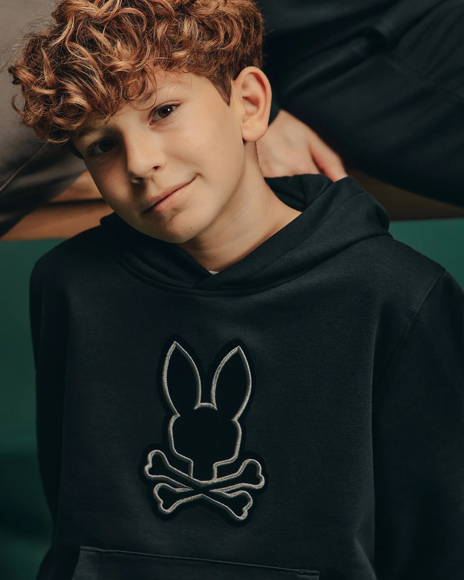 A boy with curly hair is wearing the Psycho Bunny Kids Alexander Hoodie (B0H154E200), which features a stylish bunny skull design. This versatile black hoodie perfectly complements his relaxed expression. The blurred background highlights his comfortable, laid-back fit and cool style.