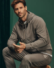 A man sits comfortably on a stool against a green background, wearing the Psycho Bunny MENS ALEXANDER HOODIE - B6H154E200 paired with matching sweatpants featuring a rabbit skull design. His short hair and beard frame his thoughtful expression as he gazes towards the camera.