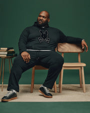A man sits confidently on a wooden chair against a dark green background. He is dressed in the BIG AND TALL ALEXANDER HOODIE - B9H154E200 from Psycho Bunny, paired with black pants and sneakers. A small stack of books rests on a side table beside him, enhancing the cozy ambiance of his comfortable hoodie ensemble.