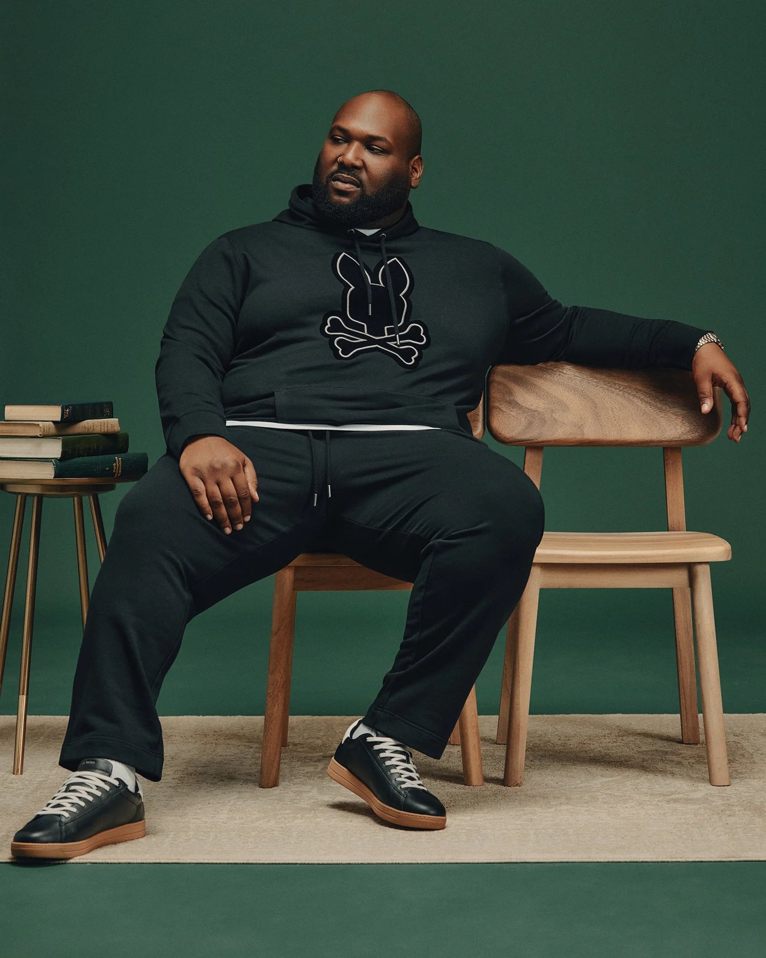 A man sits confidently on a wooden chair against a dark green background. He is dressed in the BIG AND TALL ALEXANDER HOODIE - B9H154E200 from Psycho Bunny, paired with black pants and sneakers. A small stack of books rests on a side table beside him, enhancing the cozy ambiance of his comfortable hoodie ensemble.