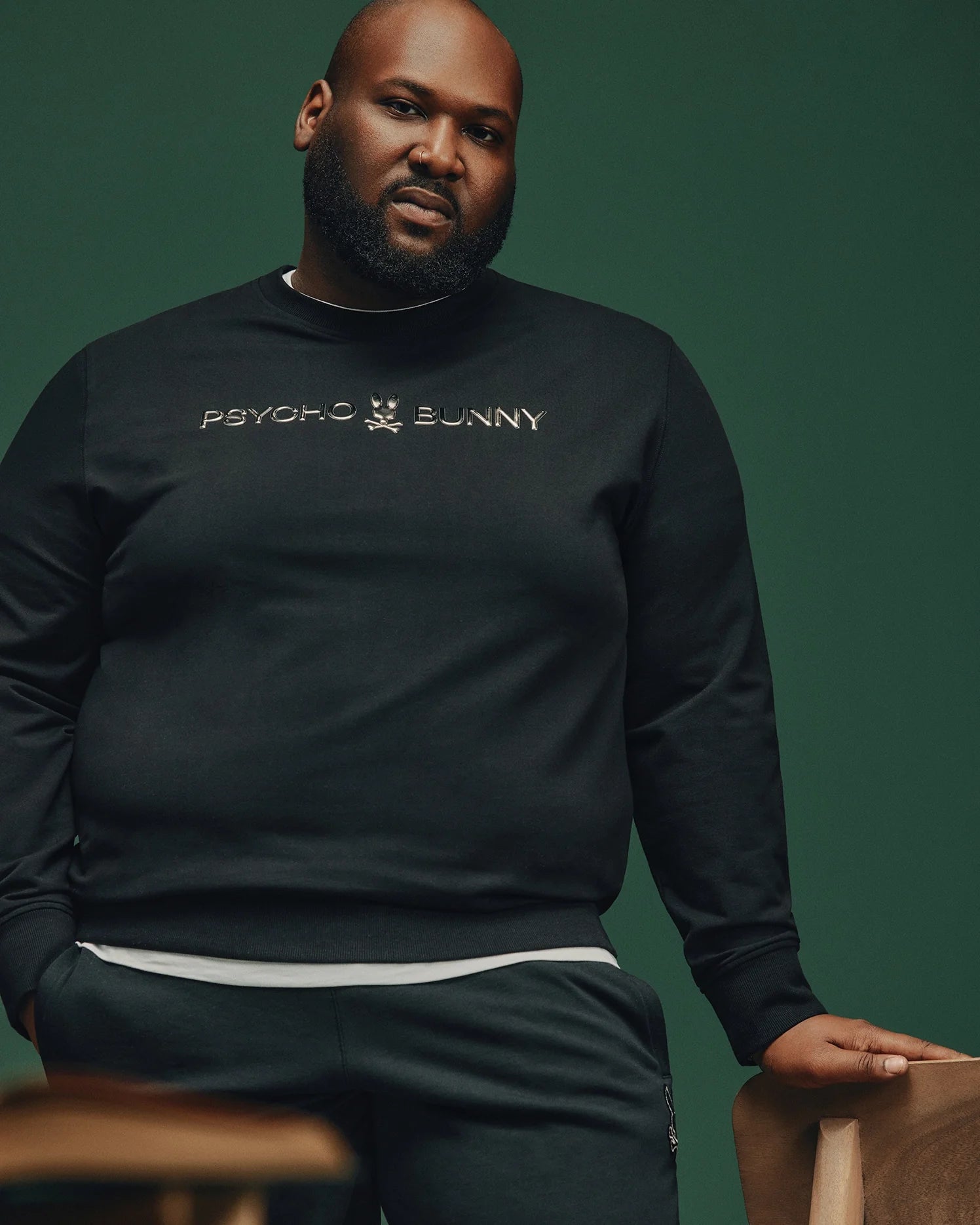 A man with a beard is wearing the BIG AND TALL LAVATA FRENCH TERRY SWEATSHIRT by Psycho Bunny. Perfect for style and comfort, he stands against a dark green backdrop, leaning casually with one hand in his pocket.
