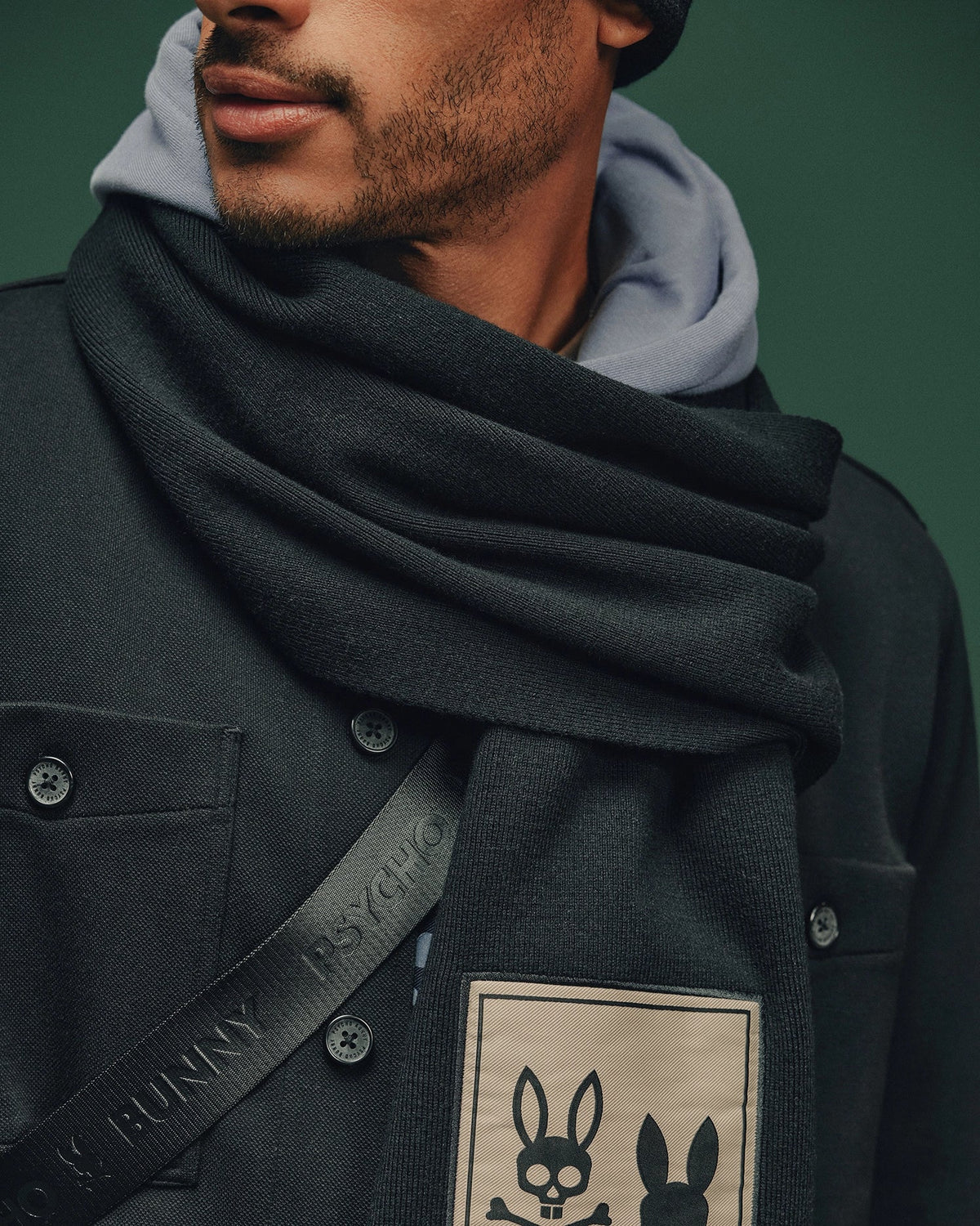 The individual wears a dark coat adorned with an elegant Psycho Bunny patch featuring two stylized bunny faces. To complement the coat, their Monroe Twill Patch Scarf by Psycho Bunny showcases a charming bunny design. Completing the ensemble are a hoodie and black beanie, all set against a vibrant green background.