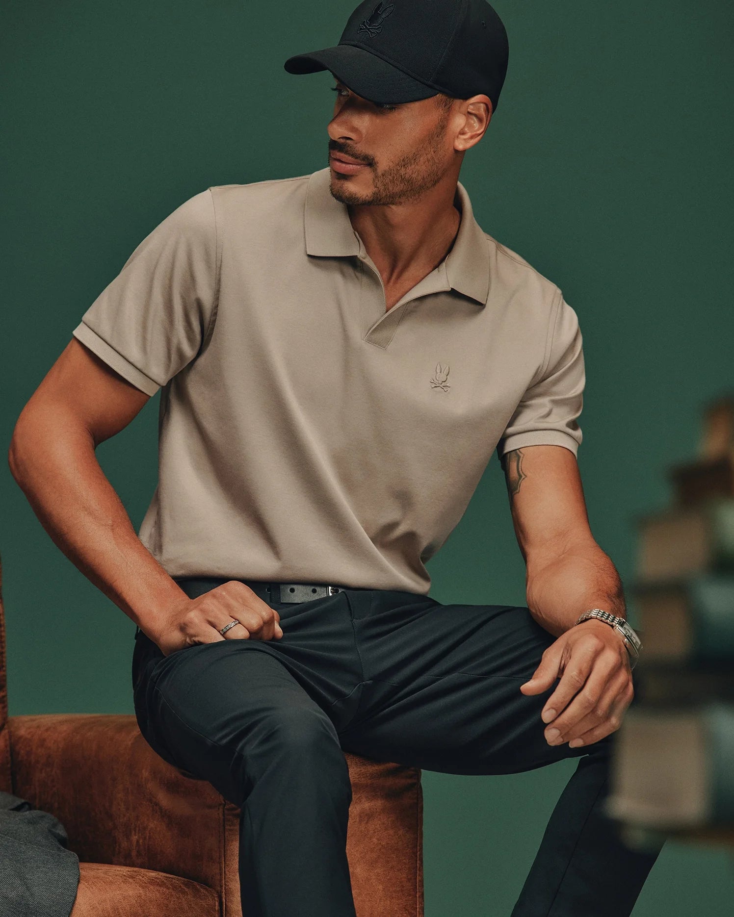 The individual, donned in a beige MENS BROADWAY PIMA INTERLOCK JOHNNY COLLAR POLO by Psycho Bunny, black pants, and a black cap, sits thoughtfully on a brown chair against a green backdrop.