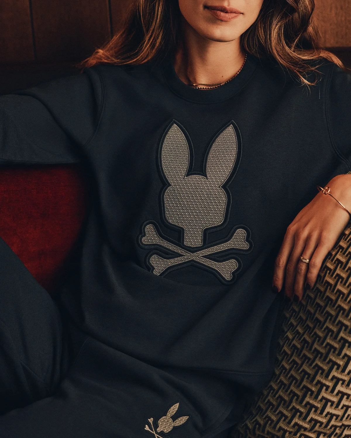 A person wearing a dark Women's Alexander Sweatshirt (B2S156E200) by Psycho Bunny, which features a prominent bunny and crossbones design, sits on a patterned chair with their face and hands partially visible. One hand rests on their knee, and the pants are adorned with an embroidered Lurex bunny design.