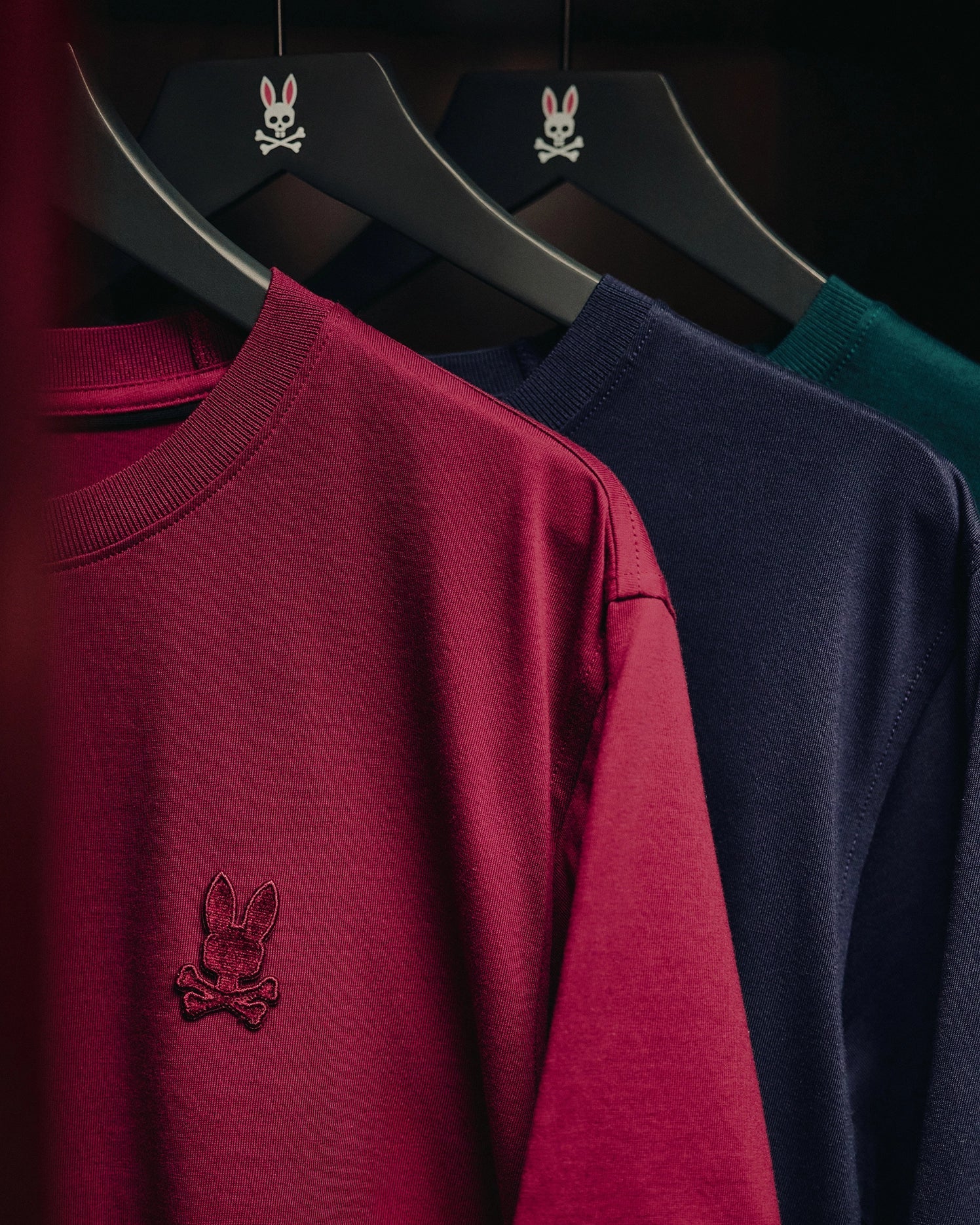 Three stylish MENS BRANSON FASHION TEEs from Psycho Bunny in burgundy, navy, and forest green hang on black hangers. Each tee features a small embroidered emblem of a rabbit with crossbones. These fashion-forward pieces are as essential to your wardrobe as any men's pima cotton tee.