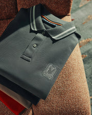 A meticulously folded MENS PIERCE PIQUE POLO from Psycho Bunny, crafted from luxurious Pima cotton, showcases an embroidered Bunny logo on the chest. This dark polo shirt features a white-striped collar and is arranged on a textured surface, gently illuminated by sunlight spotlighting its fabric.