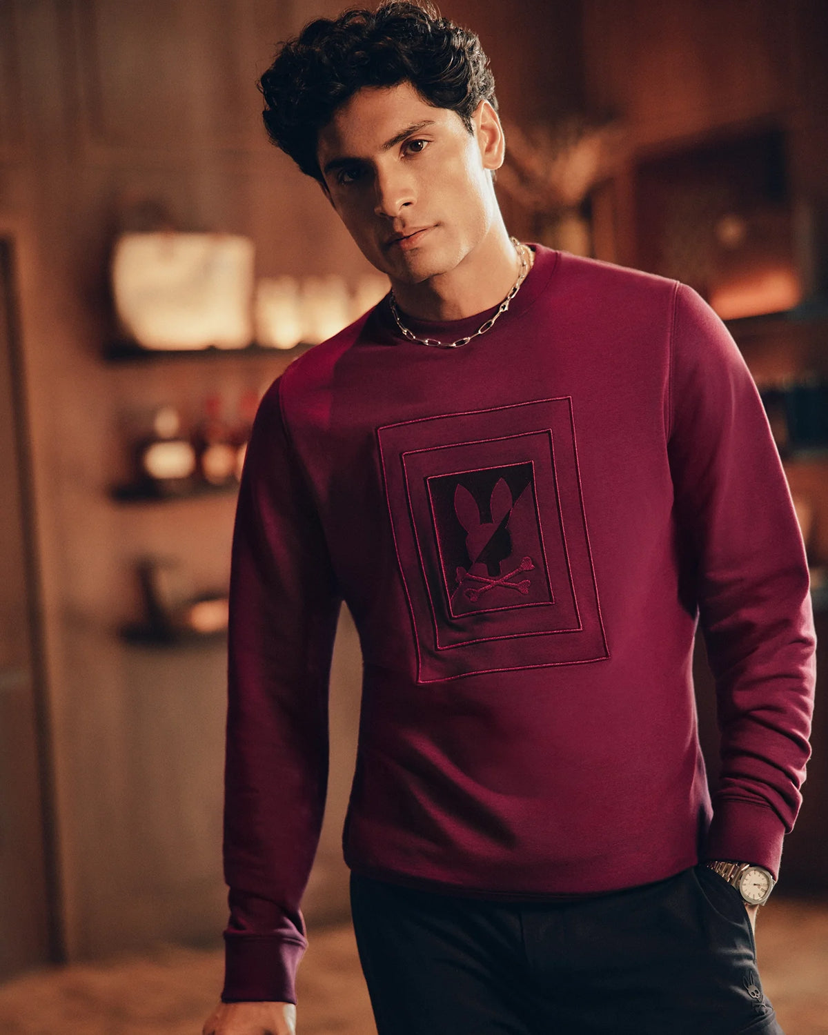 The individual stands indoors wearing a Psycho Bunny men's Oak City crew neck sweater (B6S274E200) in maroon, distinguished by its premium quality raised embroidery featuring a subtle rabbit logo. The warmly lit room highlights shelves and a plant in the blurred background as the person, with dark curly hair, gazes slightly to the side.