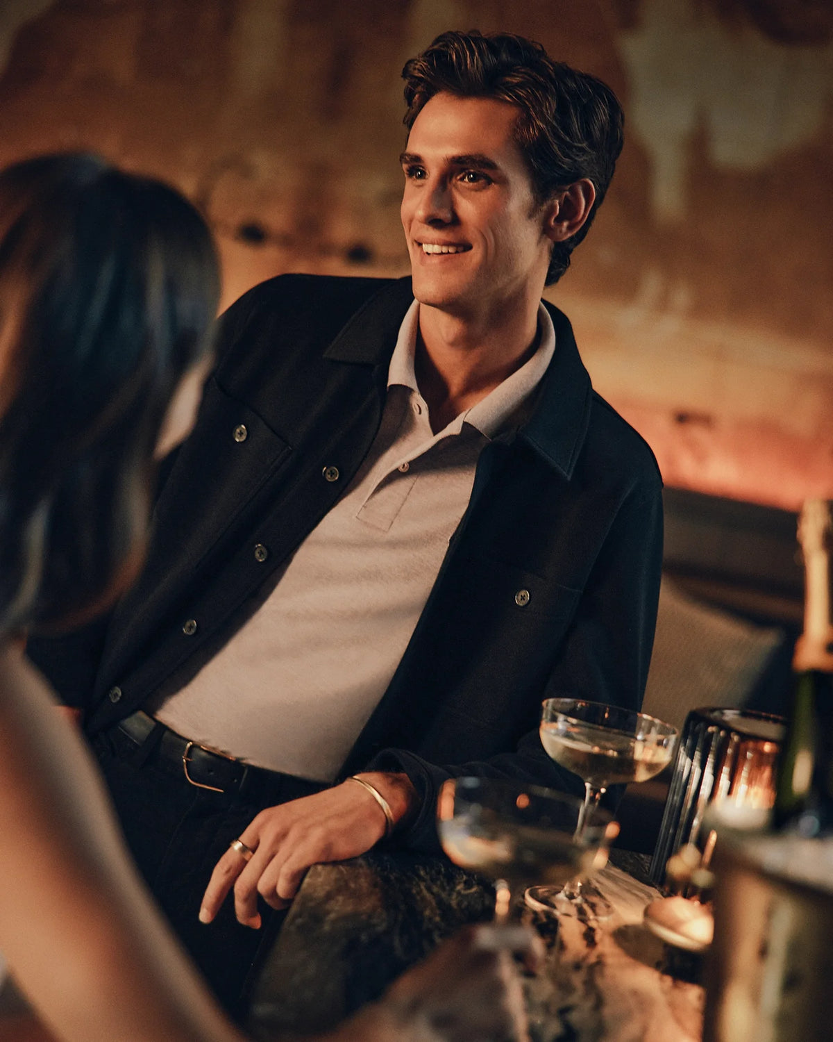 A man wearing a chic Psycho Bunny MENS BEDFORD SHIRT JACKET - B6J290B200 stands at a bar, smiling at a woman. Two glasses of champagne are next to them, enveloped in a warm ambient glow that heightens the enchanting atmosphere.