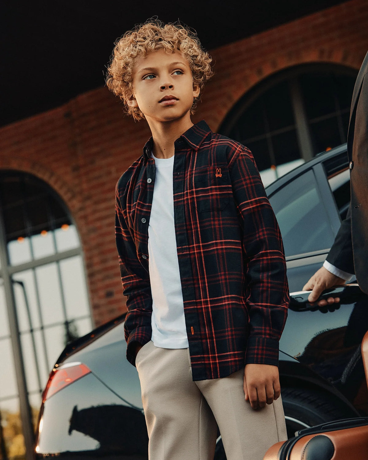A curly-haired boy stands confidently outdoors, dressed in a Psycho Bunny KIDS HAYES PLAID SHIRT - B0C842D200 in red and black over a white T-shirt, with beige pants. He is near a black car with a brick building behind him.