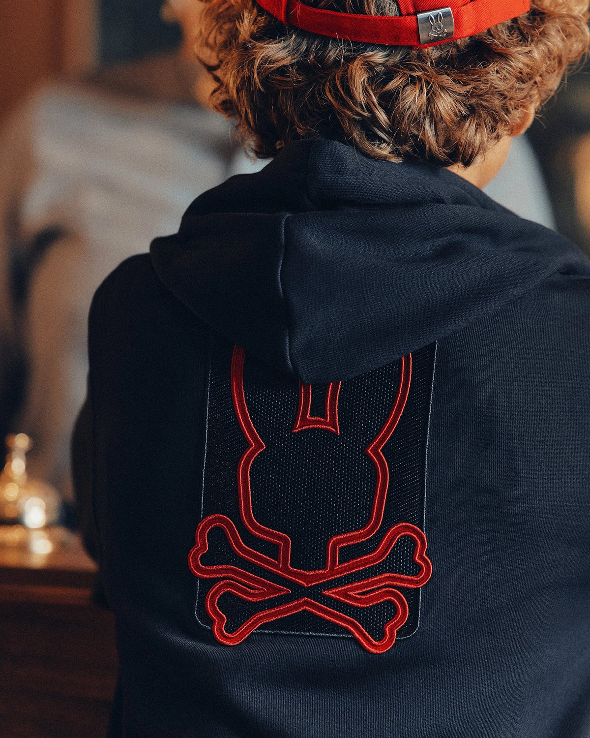 A person sports the KIDS PIERCE ESSENTIAL FRENCH TERRY ZIP HOODIE by Psycho Bunny, featuring a red-outlined stylized bunny head and crossbones design on the back. With curly hair and a red cap, they project an effortlessly cool appearance.