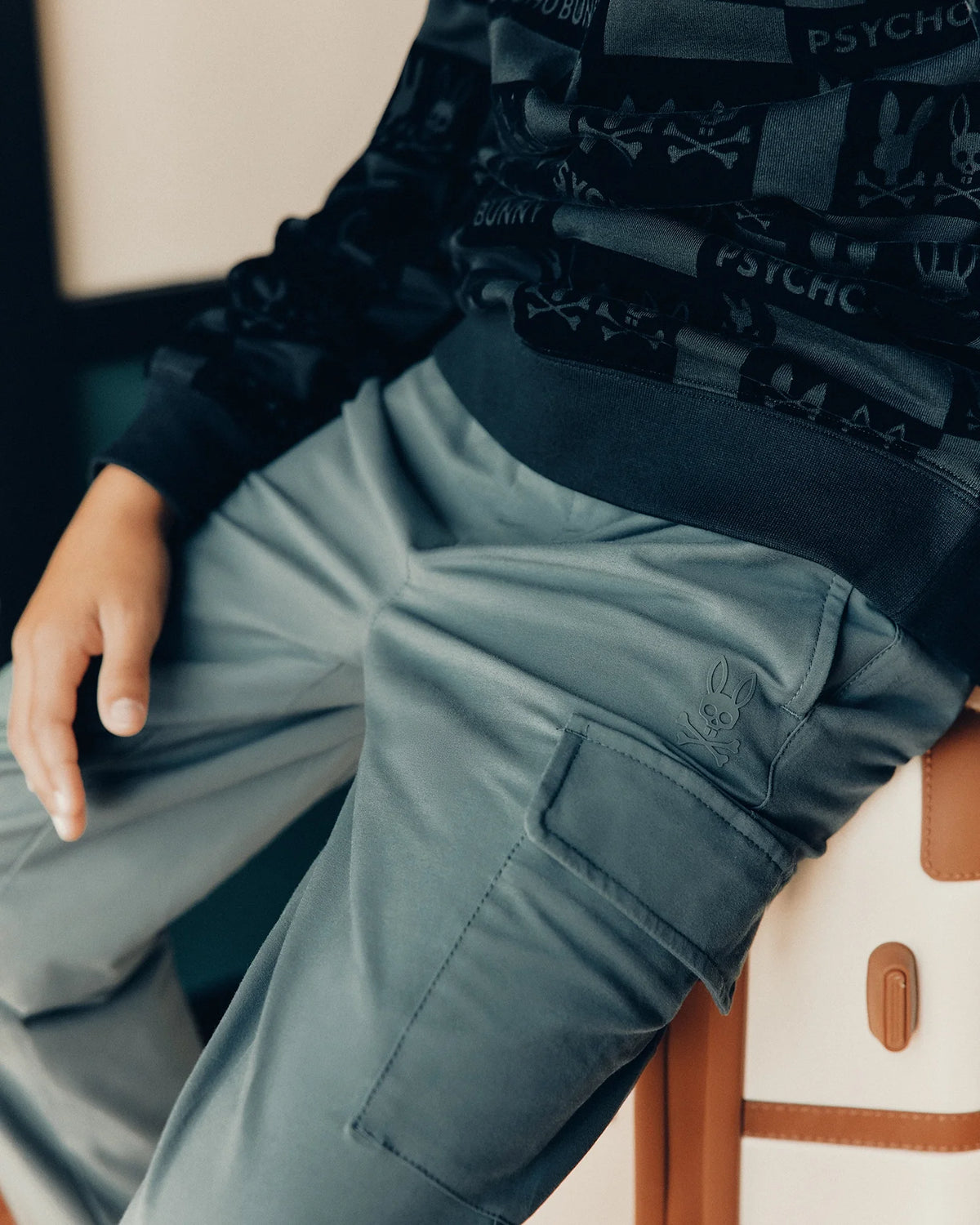 A person wearing a black sweatshirt with graphics and the trendy KIDS GREEN VALLEY CARGO PANT - B0P199E200 sits with one hand resting on their lap. The stylish trousers from Psycho Bunny feature a small rabbit logo.