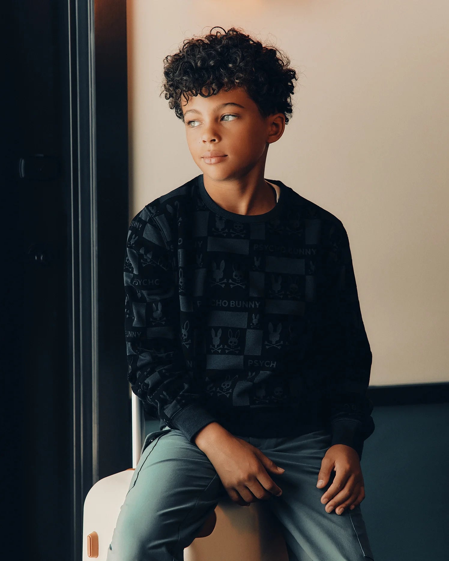 A young boy with curly hair sits on a light-colored bench, wearing the KIDS ROMEO LIGHTWEIGHT SWEATSHIRT by Psycho Bunny and light gray pants. He gazes thoughtfully to the side, surrounded by softly lit warm tones.
