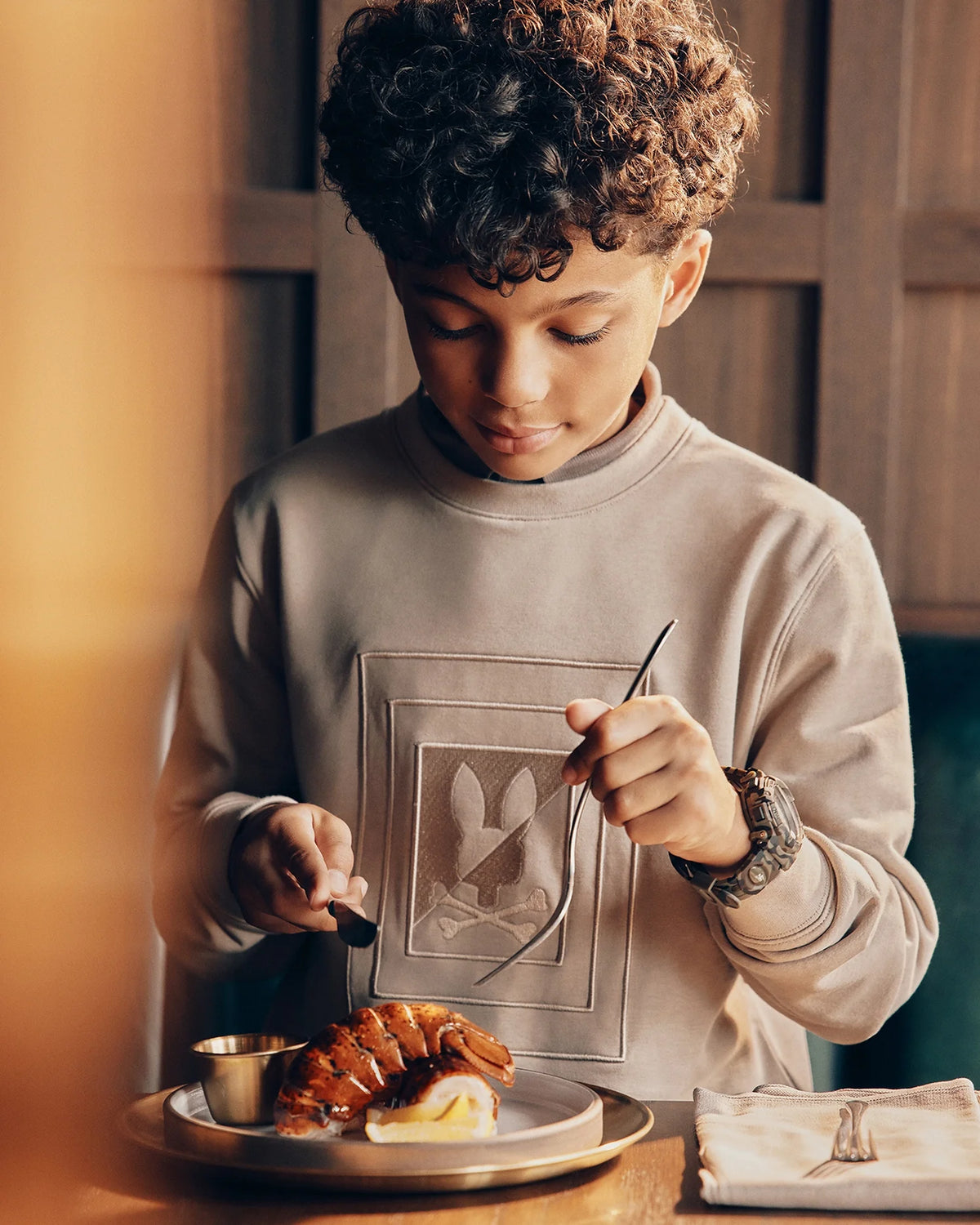 A young boy with curly hair sits at a table, using a fork and knife to cut into a croissant on his plate. The table is set with a napkin and small sauce container. He wears the KIDS OAK CITY CREW NECK SWEATSHIRT by Psycho Bunny that perfectly matches his playful spirit.