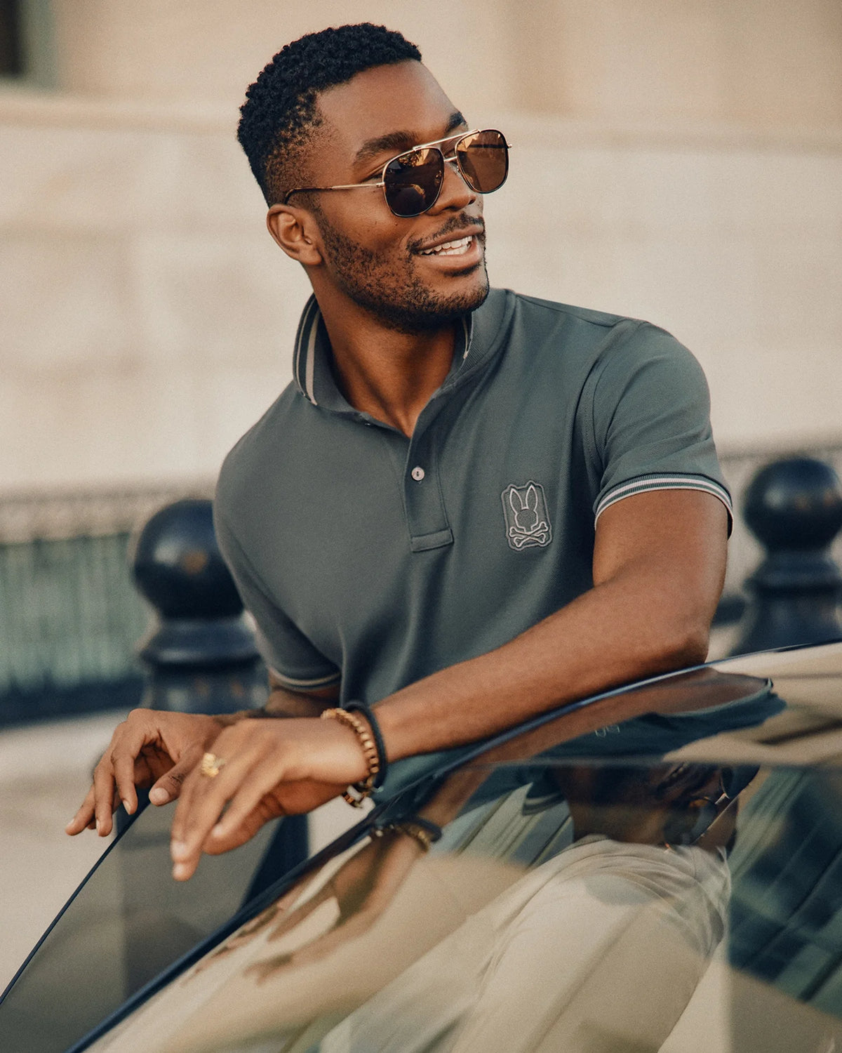 A man sporting sunglasses and a Psycho Bunny MENS PIERCE PIQUE POLO - B6K201E200, which features an embroidered bunny, leans on a car with a smile. He has short hair and a beard while wearing a bracelet on his wrist, set against an outdoor urban backdrop.