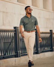 A man in sunglasses walks confidently along the pathway, wearing the green embroidered Mens Pierce Pique Polo by Psycho Bunny, expertly crafted from soft Pima cotton. He's paired it with beige pants and a brown belt, all set against a backdrop of a decorative black railing and a light-colored building.