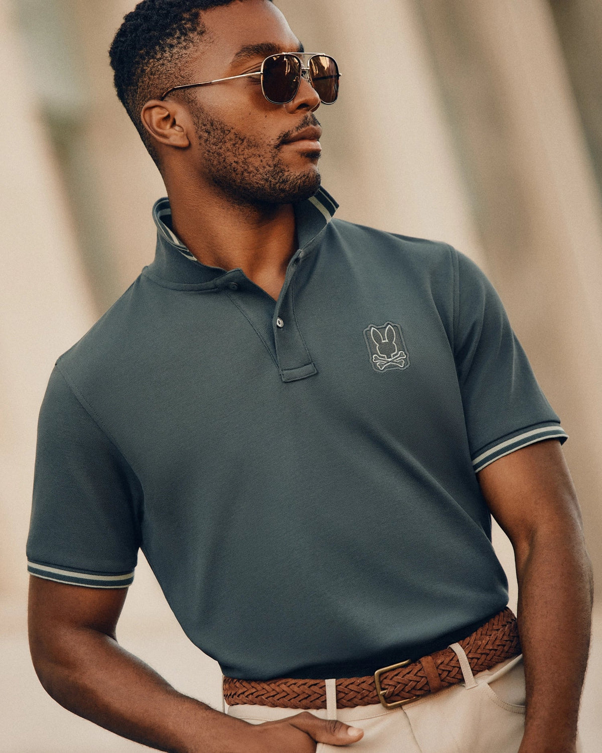 A man wearing the Psycho Bunny MENS PIERCE PIQUE POLO - B6K201E200, complete with an embroidered Bunny logo, stands confidently outdoors in sunglasses. His dark Pima cotton polo shirt is neatly tucked into beige pants, cinched by a braided brown belt. The softly blurred background highlights the man's stylish appearance.