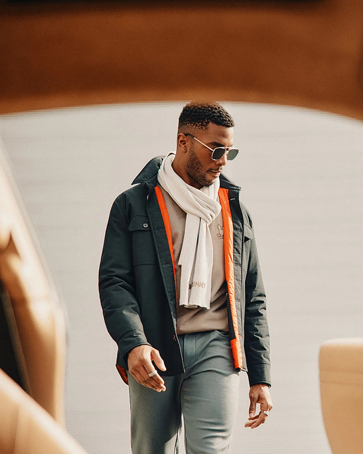A man wearing the Psycho Bunny MENS BEANIE AND SCARF BOX SET - B6X353E200, paired with sunglasses, a black jacket with an orange lining, and gray pants, walks outdoors. The background is blurred with neutral tones and some seating visible in the foreground.