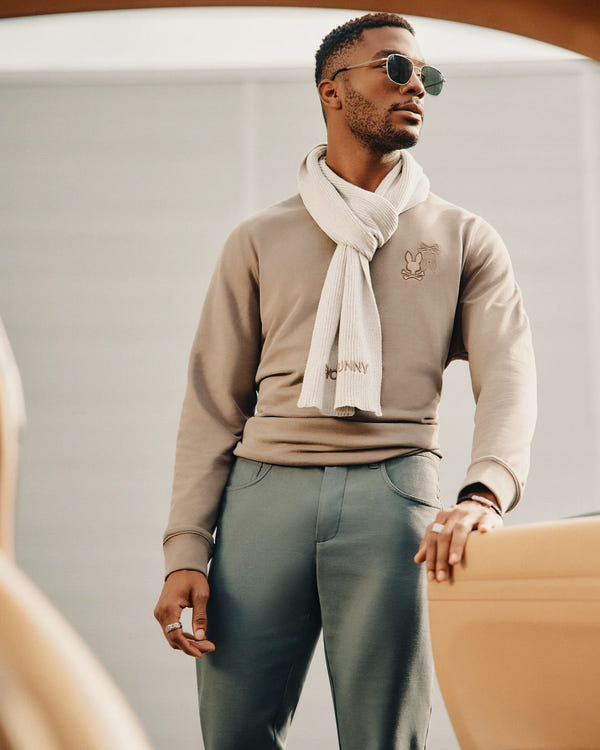 A man sporting a pair of sunglasses and the MENS BEANIE AND SCARF BOX SET by Psycho Bunny radiates confidence. He's styled in a beige sweatshirt with a subtle graphic, green trousers, and a ring, all set against a chic, modern backdrop.