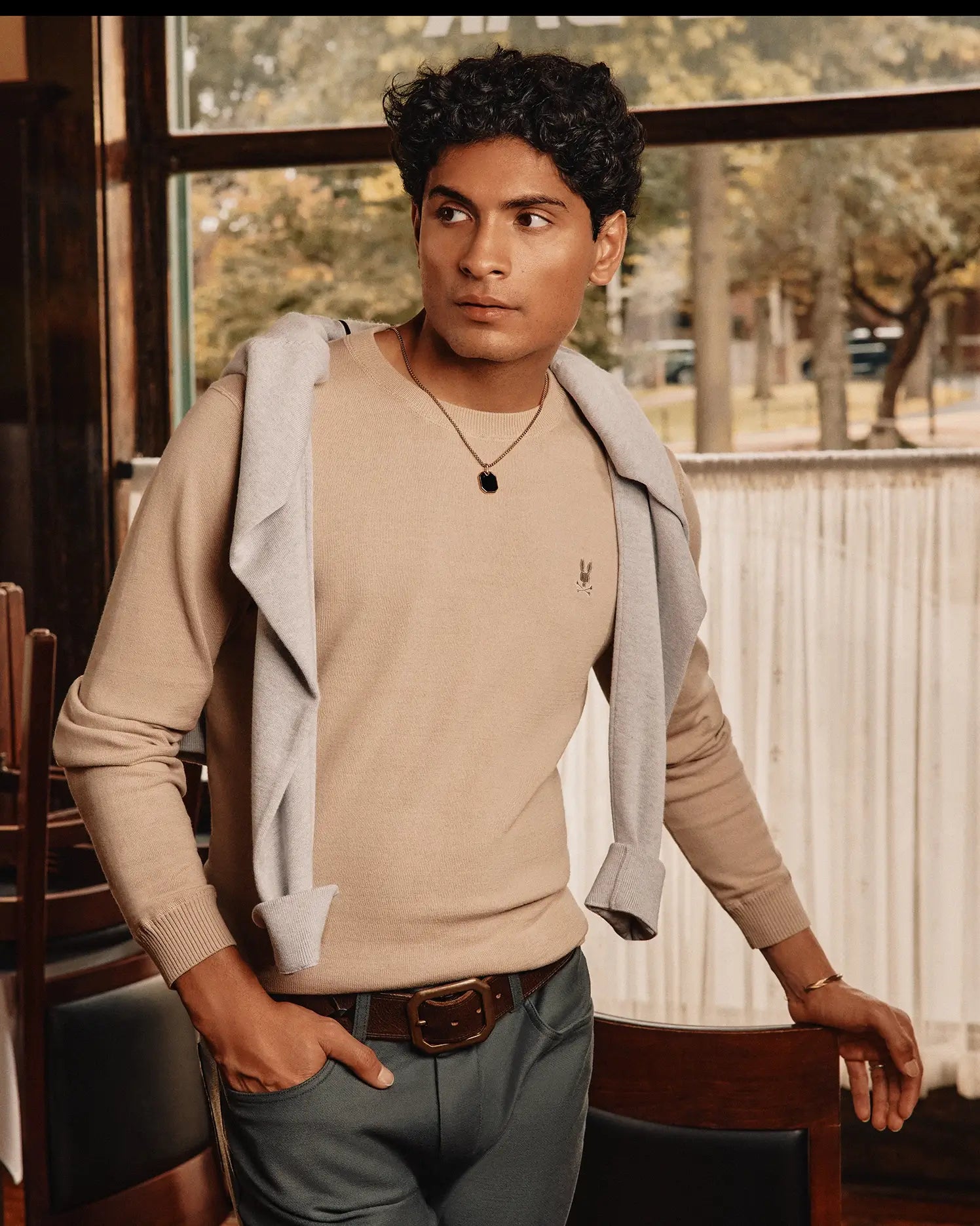 A person with curly hair stands indoors wearing a Psycho Bunny MENS BELMORE EXTRA FINE COTTON SWEATER in beige, draped with a gray sweatshirt over their shoulders and dark pants. They lean on a wooden table, looking at an embroidered Bunny logo by the window showing a blurred outdoor view.