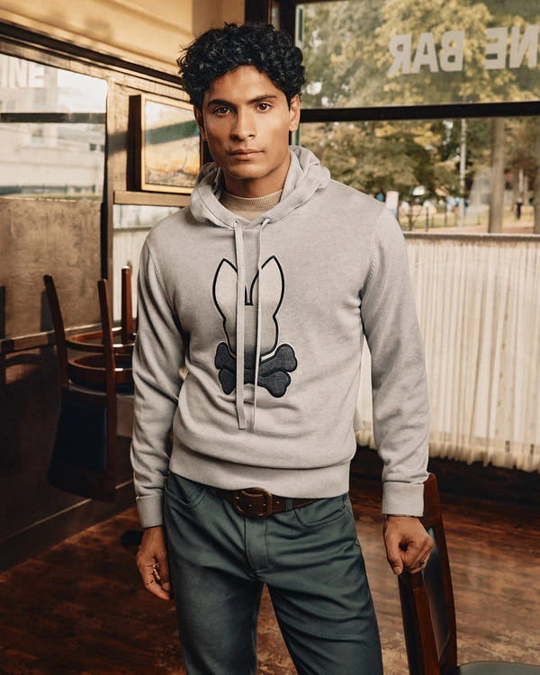 A person with dark curly hair is indoors wearing a Psycho Bunny MENS BULDER HOODED SWEATER (B6E261E200) featuring a black bunny and bone design. They pair it with dark green pants, standing in a warmly lit room with wooden chairs and large windows behind them.