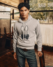 A person with dark curly hair is indoors wearing a Psycho Bunny MENS BULDER HOODED SWEATER (B6E261E200) featuring a black bunny and bone design. They pair it with dark green pants, standing in a warmly lit room with wooden chairs and large windows behind them.