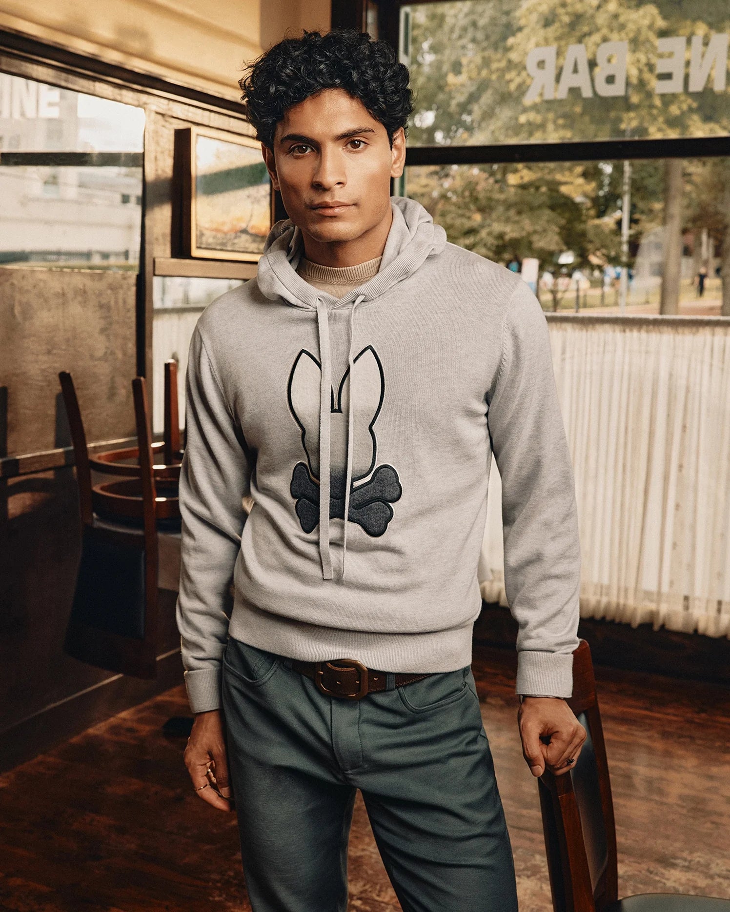 A person with dark curly hair is indoors wearing a Psycho Bunny MENS BULDER HOODED SWEATER (B6E261E200) featuring a black bunny and bone design. They pair it with dark green pants, standing in a warmly lit room with wooden chairs and large windows behind them.