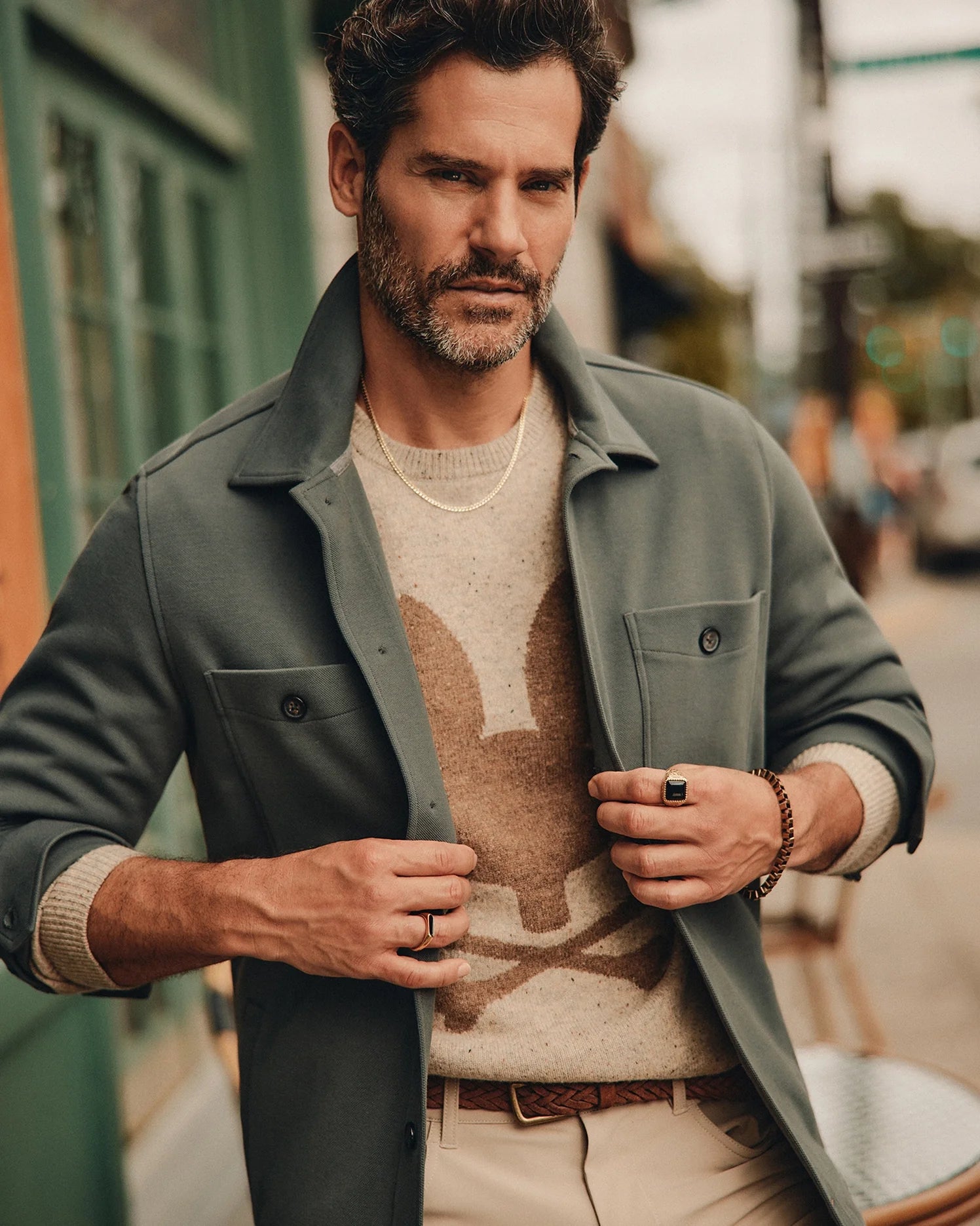 A bearded man stands confidently on a street, exuding an urban vibe. He wears the stylish Psycho Bunny Men's Bedford Shirt Jacket - B6J290B200 over a cotton-blend sweater with a rabbit design, paired with beige pants and accessories including a necklace, rings, and a bracelet. The backdrop features a green door and blurred city buildings.