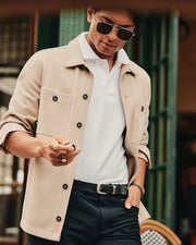 A person wearing aviator sunglasses and a beige MENS BEDFORD SHIRT JACKET - B6J290B200 by Psycho Bunny, featuring logo-engraved buttons over a white polo shirt, stands outside a cafe, looking down at something in their hand. The setting is casual and stylish.