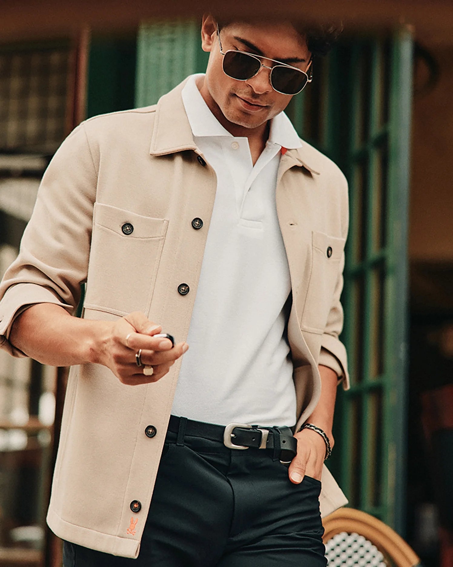 A person wearing aviator sunglasses and a beige MENS BEDFORD SHIRT JACKET - B6J290B200 by Psycho Bunny, featuring logo-engraved buttons over a white polo shirt, stands outside a cafe, looking down at something in their hand. The setting is casual and stylish.