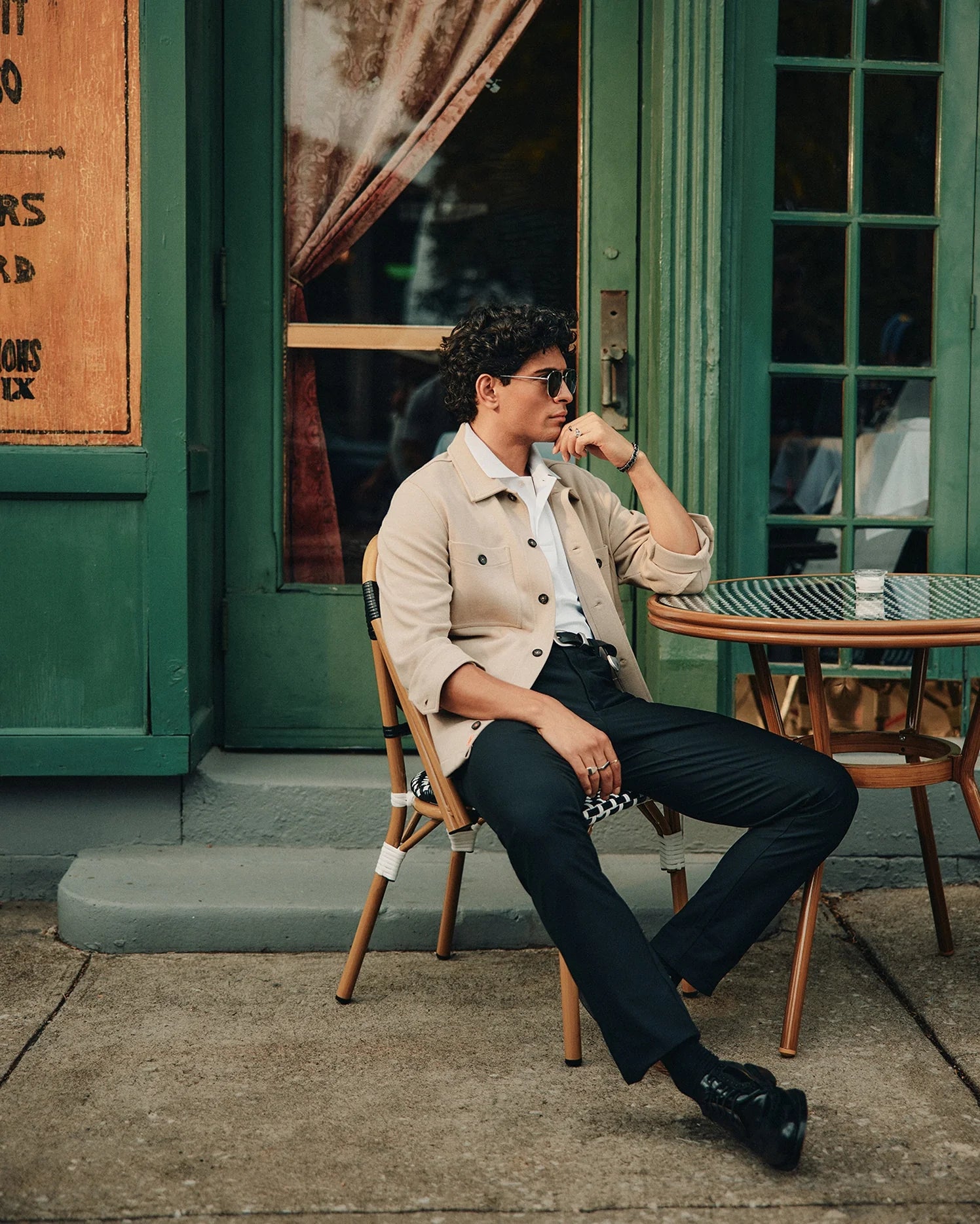 A person with curly hair sits outside a cafe on a wicker chair, leaning on one hand. They wear sunglasses, a Psycho Bunny MENS BEDFORD SHIRT JACKET - B6J290B200, a white shirt, and black trousers, their legs crossed. A small round table is next to them against the building with green doors and windows.