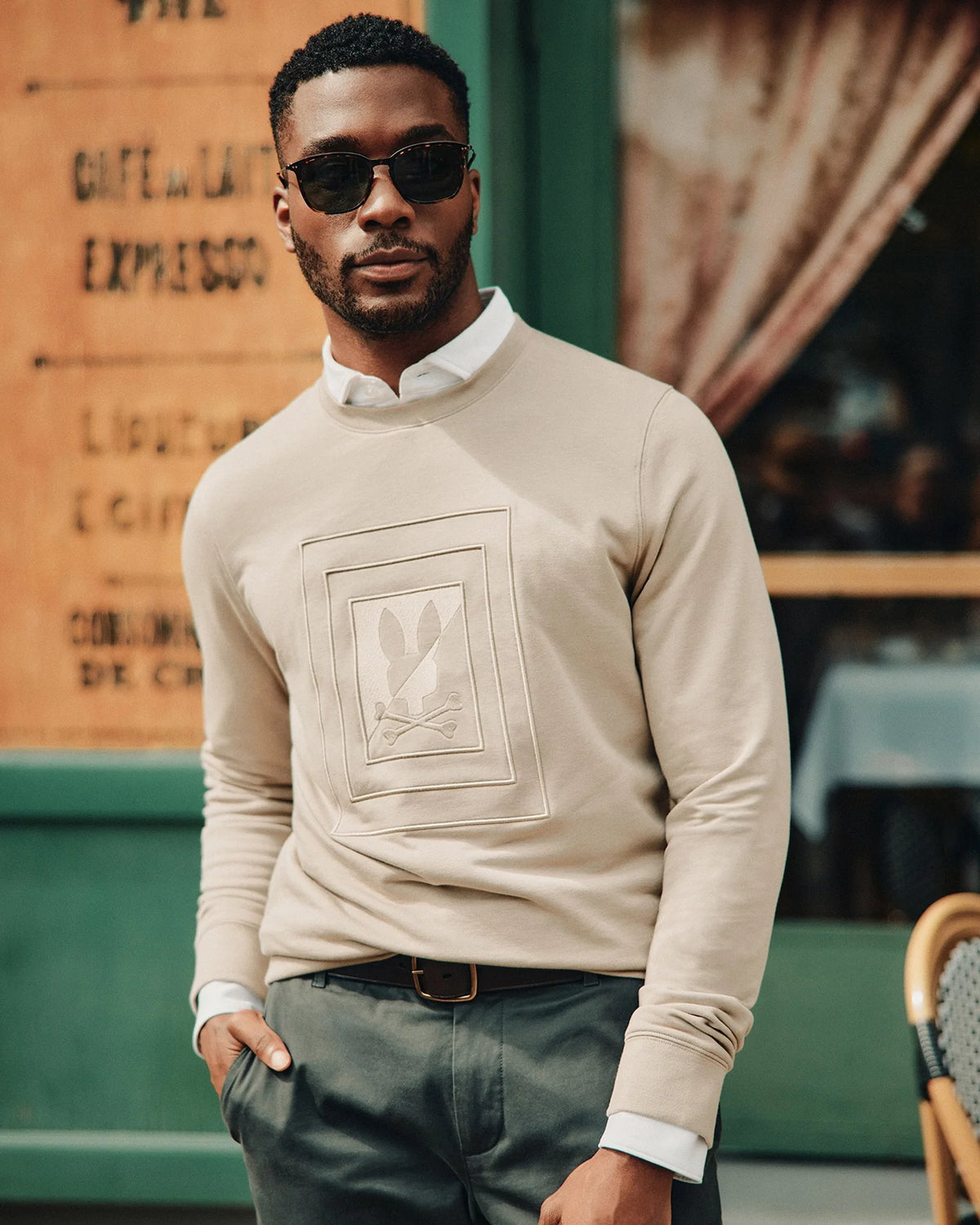 A man wearing sunglasses and the Psycho Bunny MENS OAK CITY CREW NECK (B6S274E200), showcasing a raised embroidery bunny design, stands confidently in front of a green outdoor café, styled with dark pants and a white collared shirt.