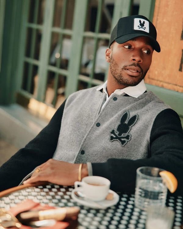 A person is seated at an outdoor cafe table, wearing an elegant Psycho Bunny MENS TOM WOOL BOMBER JACKET - B6J744D200, complemented by a white shirt and a black cap. On the table, there's a glass of water with a lemon slice and a cup of coffee. In the background is a green window.