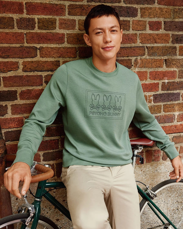 A man wearing the MENS TACOMA SWEATSHIRT - B6S927D200 by Psycho Bunny, in green, sits on a bicycle exuding luxurious comfort. He is posed against a brick wall, sporting beige pants with premium stretch and smiling at the camera.
