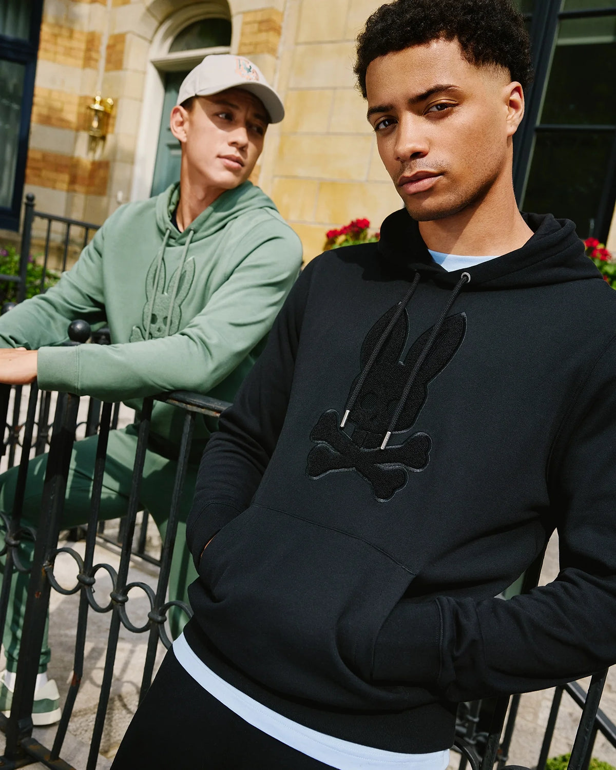 Two men stand outdoors near a wrought iron fence. One is sporting the Psycho Bunny chenille hoodie with a bunny skull design in black, while the other wears a green Men's Pacific Hoodie (B6H930D200) along with a beige cap. They appear relaxed, with a building and lush plants in the background.