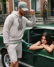 A man in a grey hoodie, Psycho Bunny MENS WALTER LIGHTWEIGHT SWEATSHORT - B6R477D200, and matching cap stands next to a parked green car. He looks down and rests one arm on the car. A woman with long dark hair leans out of the car's window, resting her arms and chin on the edge of the window frame.