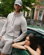 A man in a grey hoodie made of high-quality cotton blend, wearing Psycho Bunny's MENS WALTER LIGHTWEIGHT SWEATSHORT - B6R477D200, and a baseball cap with a white skull and crossbones emblem stands by a green car. A woman with long dark hair leans out of the car's window, resting her arms on the door. They are on a residential street with trees and buildings.