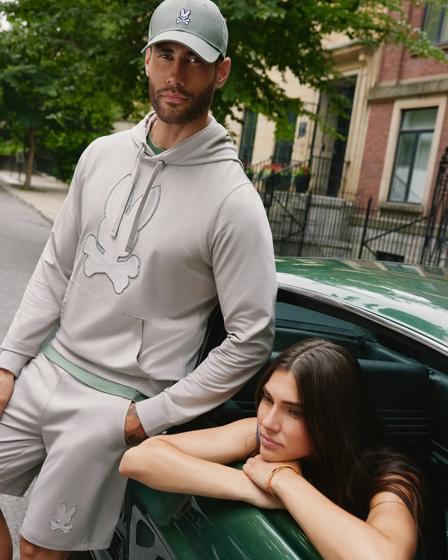 A man in a grey hoodie made of high-quality cotton blend, wearing Psycho Bunny's MENS WALTER LIGHTWEIGHT SWEATSHORT - B6R477D200, and a baseball cap with a white skull and crossbones emblem stands by a green car. A woman with long dark hair leans out of the car's window, resting her arms on the door. They are on a residential street with trees and buildings.