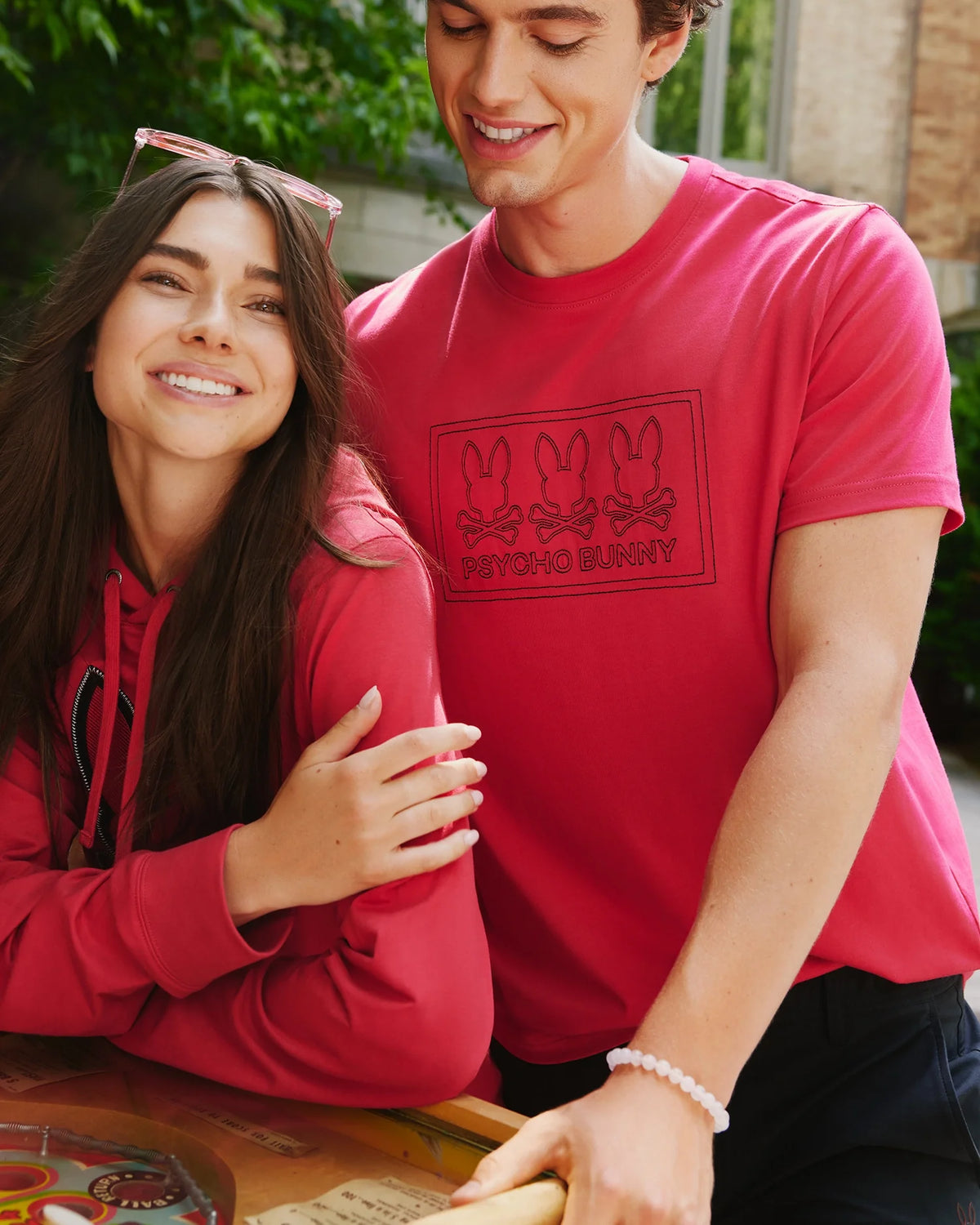 A smiling woman with long hair and sunglasses rests her arm on a man who is wearing a red shirt from Psycho Bunny's collection. They are outdoors near a building, enjoying a relaxed moment together.