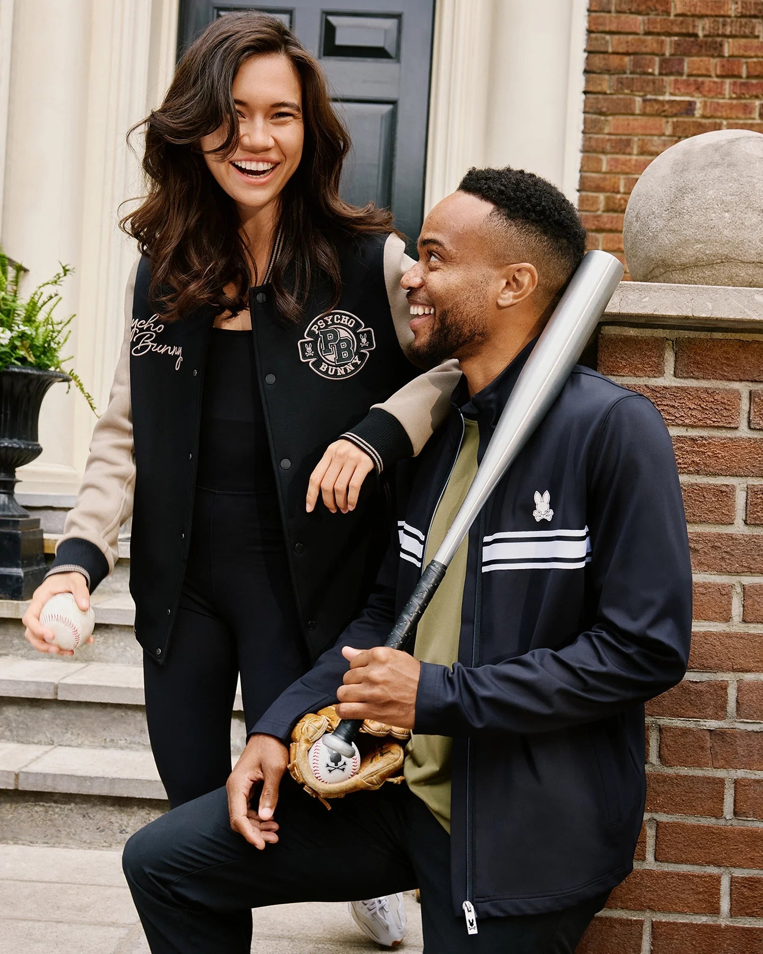 A woman and a man pose on steps. The woman, smiling, holds a baseball. The man, also smiling, leans on a baseball bat with a glove on the other hand. They both wear stylish track jackets; the man is wearing the Psycho Bunny MENS TERRELL SPORT ZIP UP JACKET - B6J643C200. A brick wall and a black door are in the background.
