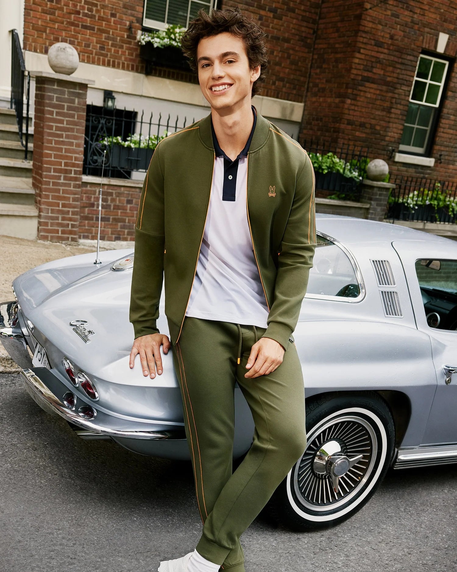 A young man with short, dark hair smiles while leaning against a vintage silver car parked on a residential street. He is wearing the Psycho Bunny MENS TRAVIS TRACK PANT - B6P143D200 in olive green made from high-quality cotton-blend fabric, paired with a white and black polo shirt and white sneakers. A brick building with a window and steps is in the background.