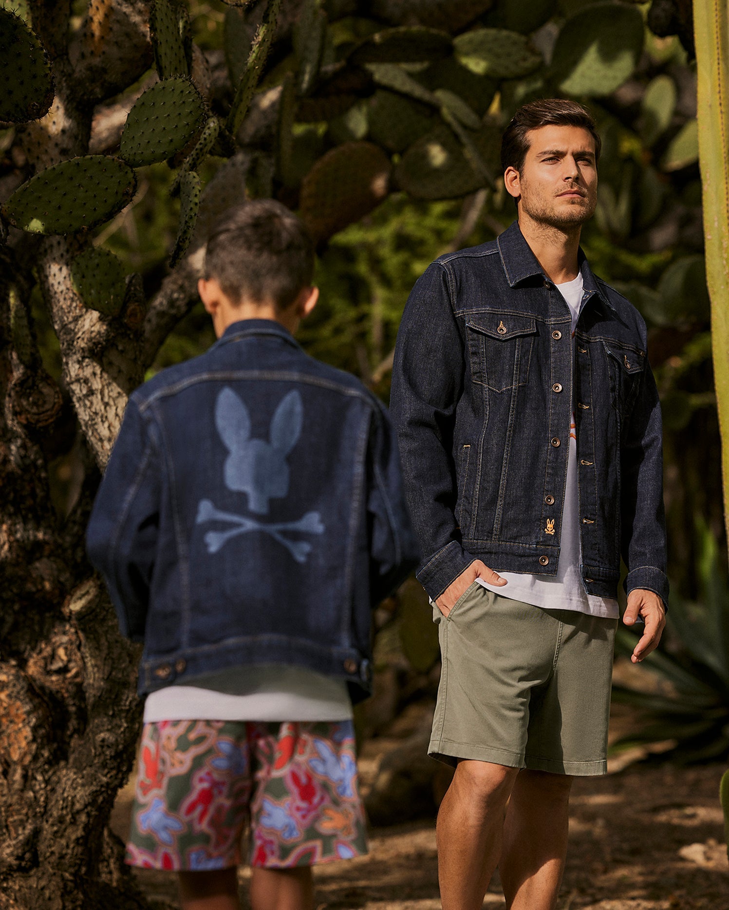 Two people are outdoors surrounded by cactus plants. One faces the camera showcasing men's fashion in the MENS PATHE DENIM JACKET - B6J741X1CO by Psycho Bunny and shorts, while the other, with their back turned, sports raw denim featuring a skull design. They stand on a dirt path.