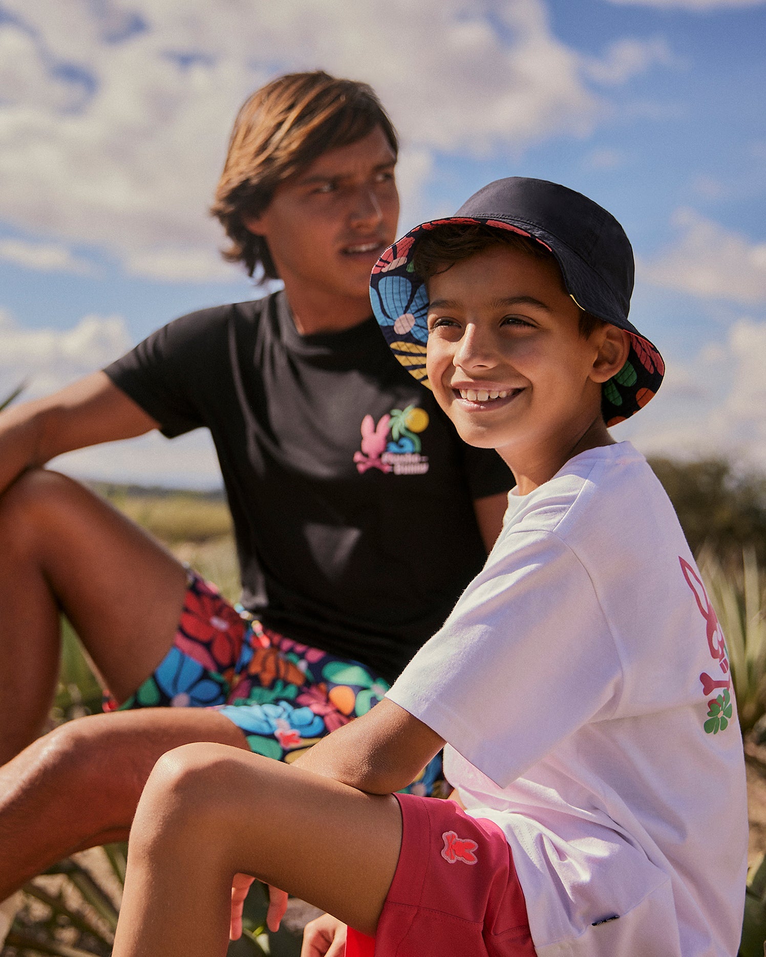 A young person wearing a colorful bucket hat and a white t-shirt sits beside an older individual in a black t-shirt with Psycho Bunny MENS MENTZ SWIM TRUNK - B6W320B2SW. Both are outdoors with a bright sky in the background. The young person is smiling while the older individual looks off to the side.